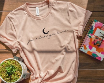 May include: A light peach-colored crew neck t-shirt featuring a black crescent moon icon and the black cursive text "your design here" printed on the chest. The shirt has short sleeves and is gathered in a knot at the bottom hem.