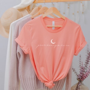 May include: A coral-coloured short-sleeve t-shirt with a crew neckline and rolled sleeves. The front features a white graphic of a crescent moon above the cursive text "your design here". The shirt is displayed on a wooden hanger.