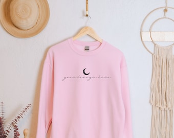 Pode incluir: Uma camisola de gola redonda em rosa claro com um gráfico de lua crescente preta e o texto "your design here" em letra cursiva. A peça está pendurada num cabide de madeira, apresentando mangas compridas e uma gola canelada.