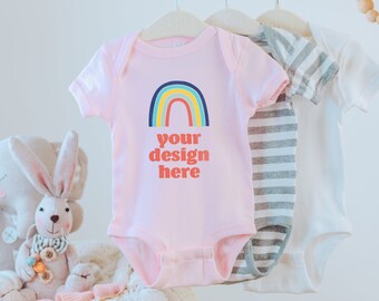 May include: A light pink baby bodysuit featuring a colorful rainbow graphic and the text your design here in red letters. The garment hangs on a hanger alongside a grey and white striped bodysuit and a plain white bodysuit on a wooden rack.