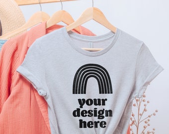 May include: A light gray short-sleeved t-shirt featuring a black graphic of a rainbow arch above the text "your design here" in a bold, rounded font. The shirt is tied in a knot at the bottom center and hangs on a wooden hanger.