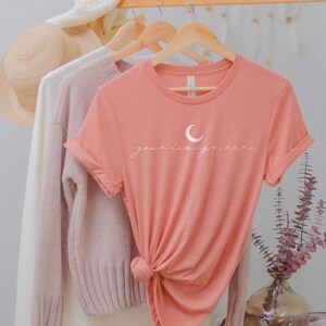 May include: A peach-coloured short-sleeve t-shirt featuring a white crescent moon graphic and the text "your design here" in a cursive font. The shirt is tied in a knot at the bottom and hangs on a wooden hanger alongside other garments.