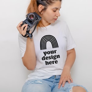 May include: A white short-sleeved t-shirt featuring a black graphic of a rainbow arch above the text "your design here" in a bold, rounded font. The shirt is paired with light blue denim jeans that have distressed detailing on the thighs.