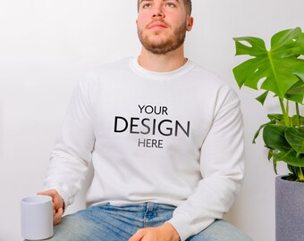 May include: A white crewneck sweatshirt featuring the black text YOUR DESIGN HERE printed on the chest. The sweatshirt is paired with blue denim jeans, and a plain white ceramic mug is held in one hand, with a green monstera plant nearby.