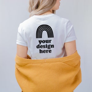 May include: A white short-sleeved t-shirt featuring a black graphic of a rainbow and the text "your design here" printed on the back. The t-shirt is worn under a mustard yellow ribbed cardigan that is draped loosely around the lower back area.