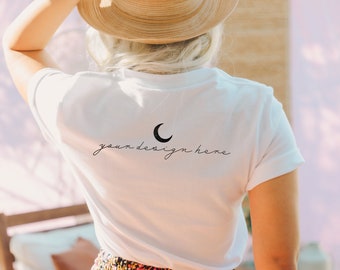 May include: A white short-sleeved t-shirt featuring a black crescent moon graphic and the text "your design here" in a cursive script font printed on the back. The shirt is worn with a tan straw hat and a patterned skirt with floral details.