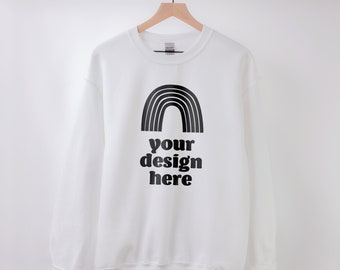 May include: A white long-sleeve crewneck sweatshirt featuring a black graphic of a rainbow arch positioned above the black text that reads "your design here". The garment is displayed on a wooden hanger against a plain, light-coloured background.