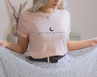 May include: A light pink short-sleeved t-shirt featuring a black crescent moon graphic and the text "your design here" in a cursive font. The shirt has a crew neckline and rolled sleeves, paired with a black belt and a white skirt with black dots.