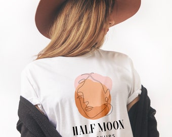 May include: A white t-shirt featuring a graphic of a single-line drawing of a face overlaid on pink and orange abstract shapes. Below the graphic, the text reads HALF MOON MOCKUPS in a black serif font. The shirt is worn with a brown hat and black cardigan.