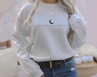 May include: A white long-sleeve sweatshirt featuring a black crescent moon graphic and the text "your design here" in a cursive script font centred on the chest. The sweatshirt has a crew neckline and is paired with blue denim jeans and a black belt.