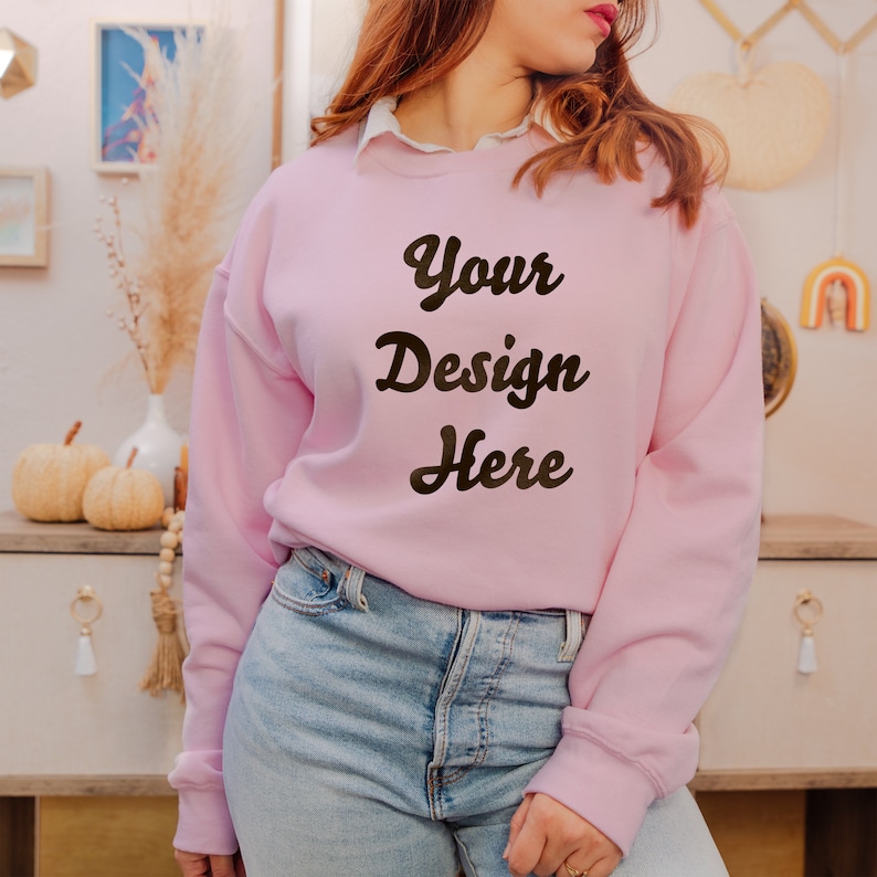 May include: A light pink long-sleeve crewneck sweatshirt featuring the dark brown text Your Design Here printed on the front. The sweatshirt is worn over a white collared shirt and paired with light blue denim jeans, creating a casual look.