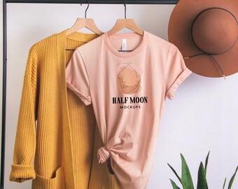 May include: A peach-colored short-sleeve t-shirt with a black line art face graphic and the text HALF MOON MOCKUPS, displayed on a hanger next to a mustard yellow open-front cardigan and a brown wide-brimmed hat on a black metal clothing rack.