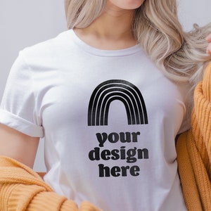 May include: A white short-sleeved t-shirt featuring a black graphic of a rainbow arch above the text "your design here" in a bold, rounded font. The shirt has a crew neckline and rolled sleeves, layered under a mustard yellow knit cardigan.