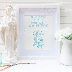 May include: White picture frame mockup with a white mat opening, 8x10 inches. The frame is on a white surface with a white statue of the Virgin Mary and a white pitcher with white flowers.