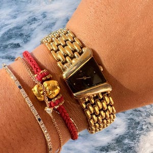 May include: A gold watch with a black rectangular face and a linked strap is shown. Beside the watch are three bracelets: a delicate chain with small blue stones, a red braided bracelet with a gold charm, and a thin gold chain.