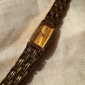 Puede incluir: Un reloj Seiko dorado con una esfera rectangular y una correa de eslabones a juego. La esfera del reloj es dorada con manecillas e indicadores de hora negros. El reloj se muestra sobre una superficie texturizada de color blanquecino.