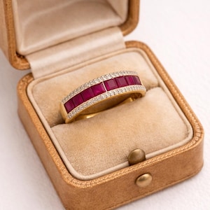 Pode incluir: Um anel de banda de ouro com uma fila de cinco rubis vermelho-escuro de corte quadrado dispostos horizontalmente no centro, ladeados por duas filas de pequenos diamantes brancos em pavé nas bordas, dentro de uma caixa de joias bege.