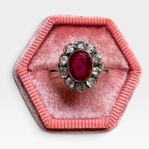 May include: A silver-toned ring featuring a central oval-cut deep red gemstone surrounded by a halo of ten small, clear, round-cut stones. The ring is displayed centered within a hexagonal, pink velvet-lined jewelry box with ribbed edges.