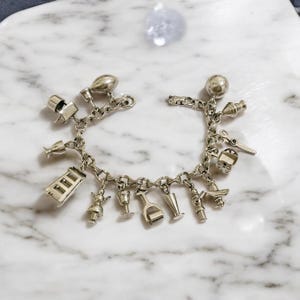 Könnte beinhalten: Ein silbernes Charm-Armband mit einer Vielzahl von Charms, darunter ein Haus, ein Weinglas, ein Schuh, eine Glocke und eine Schere. Das Armband liegt auf einer weißen Marmorfläche.