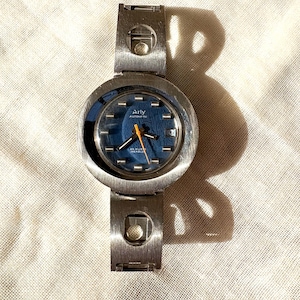 May include: A vintage silver-toned watch with a blue face and orange hands. The watch has the word "Arly" printed on the face. The watch band is made of silver-toned metal. The watch is displayed on a white, textured surface.