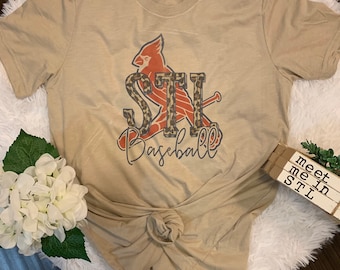 May include: A tan t-shirt with a baseball themed graphic featuring a red cardinal, the letters "STL" and the word "Baseball". The graphic is in a leopard print pattern.
