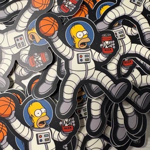 May include: A collection of stickers featuring Homer Simpson in a white astronaut suit. Homer is holding a basketball and a can of Duff Beer. The stickers have a black outline and a space-themed background.