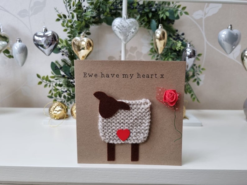 May include: A brown card with the text "Ewe have my heart x" and a knitted sheep with a red heart on its chest. A red rose with a green stem is on the right side of the card.