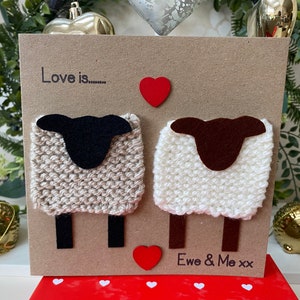 May include: A handmade card with a brown background featuring two knitted sheep with felt heads and legs. The card says "Love is..." with a red heart above the sheep and "Ewe & Me xx" below the sheep.