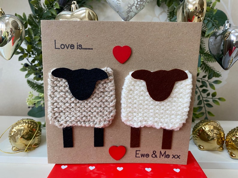 May include: A handmade card with a brown background featuring two knitted sheep with felt heads and legs. The card says "Love is..." with a red heart above the sheep and "Ewe & Me xx" below the sheep.