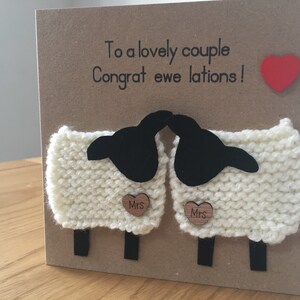 May include: A card with a punny message "To a lovely couple, Congrat ewe lations!"  Two white knitted sheep with black faces are holding a heart-shaped wooden sign that says "Mrs". A red heart is in the top right corner.