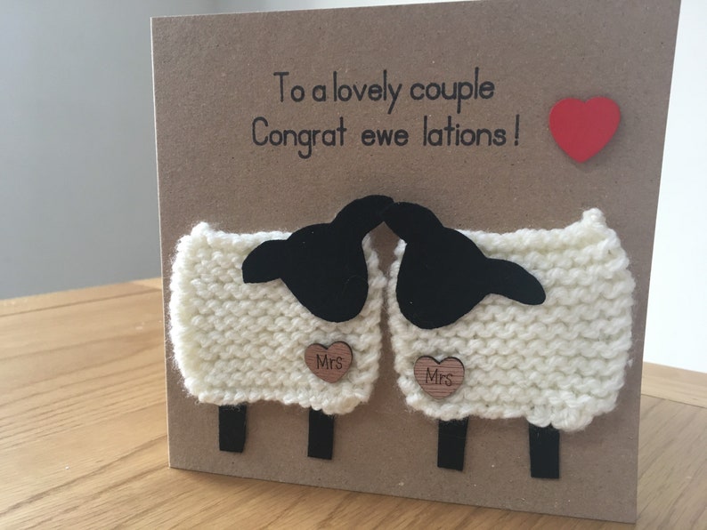 May include: A card with a punny message "To a lovely couple, Congrat ewe lations!"  Two white knitted sheep with black faces are holding a heart-shaped wooden sign that says "Mrs". A red heart is in the top right corner.