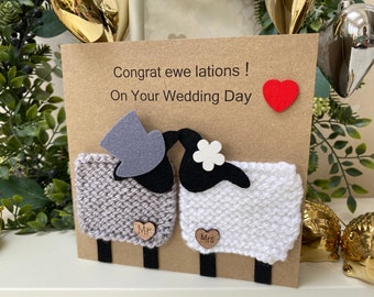 May include: A brown card with the text "Congrat ewe lations! On Your Wedding Day" and a red heart. The card features two knitted sheep, one gray and one white, with a black felt top hat on the gray sheep and a white felt flower on the white sheep. The sheep are kissing.