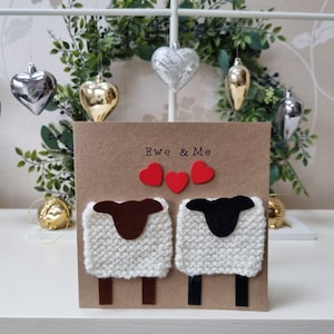 May include: A brown card with the text "Ewe & Me" and two white knitted sheep with brown and black faces. The sheep are holding red hearts.