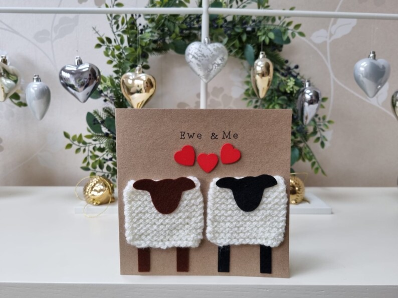 May include: A brown card with the text "Ewe & Me" and two white knitted sheep with brown and black faces. The sheep are holding red hearts.