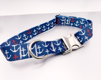 May include: A blue fabric collar featuring a repeating pattern of white and red anchors. The collar includes a silver-toned metal side-release buckle and a matching silver-toned metal D-ring for attachment, providing a secure and adjustable fit.