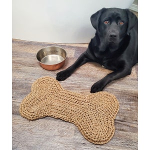 May include: A woven, bone-shaped dog bed in a natural tan colour. A copper-coloured dog bowl sits nearby. A black Labrador Retriever is lying down next to the bed, looking at the camera.