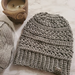 May include: A gray crocheted beanie with a ribbed brim and a textured stitch pattern. A small tag with the letters "RXR" is attached to the brim. A ball of gray yarn and a brown owl-shaped candle holder are also in the image.