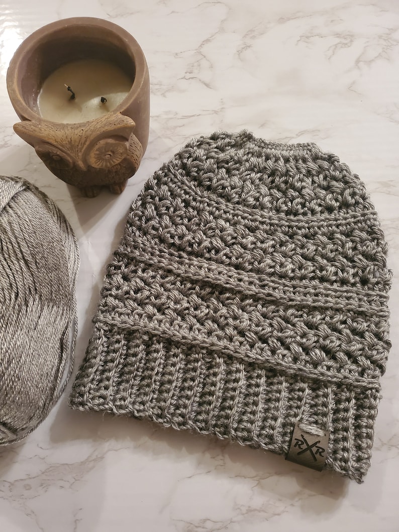May include: A gray crocheted beanie with a ribbed brim and a textured stitch pattern. A small tag with the letters "RXR" is attached to the brim. A ball of gray yarn and a brown owl-shaped candle holder are also in the image.