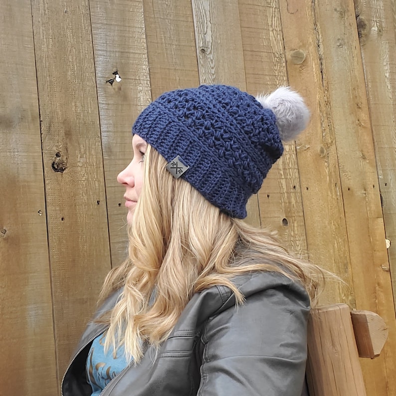 May include: A navy blue crocheted beanie with a gray faux fur pom-pom. The hat features a textured stitch pattern and a small rectangular tag with a logo. The hat is designed for cold weather.