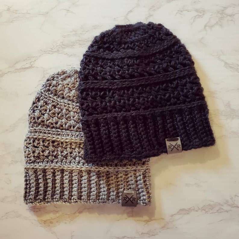 May include: Two handmade crochet beanies in the image. One is a dark charcoal gray, the other is a lighter gray with a textured stitch pattern. Both beanies have a ribbed brim and a small, rectangular tag with a logo.