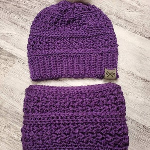 Puede incluir: Un conjunto a juego de gorro y braga de cuello de ganchillo morados. El gorro tiene un pompón de piel sintética gris y una pequeña etiqueta con las letras "RXR". El conjunto está hecho de un patrón de puntada texturizada.