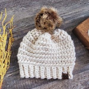 Puede incluir: Gorro de punto crema hecho a mano con un pompón de piel sintética marrón. El gorro presenta un borde acanalado y un patrón de puntada texturizada. Una pequeña etiqueta de cuero marrón con un logotipo está cosida en el borde. Accesorio de invierno.
