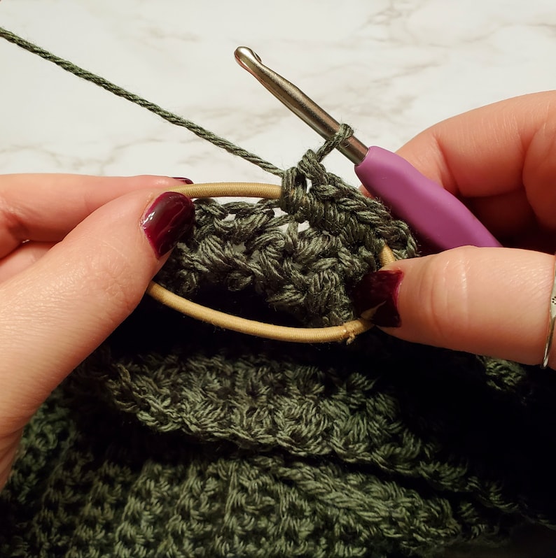 May include: Close-up of crochet work in progress. A green crocheted fabric is held with a hair tie. A crochet hook with a purple handle is inserted into the fabric, with green yarn attached. The image shows the beginning stages of a crochet project.