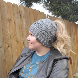 May include: A gray crocheted beanie with a ponytail opening. The hat has a textured stitch pattern and a ribbed brim. A small tag with an "X" logo is sewn on the side. The person is wearing a blue shirt and a gray leather jacket.