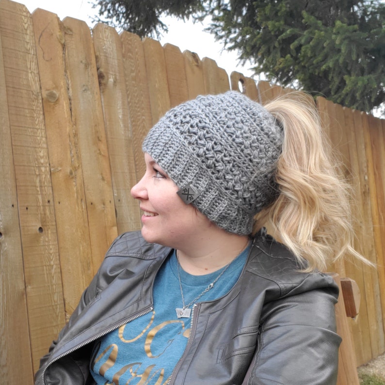 May include: A gray crocheted beanie with a ponytail opening. The hat has a textured stitch pattern and a ribbed brim. A small tag with an "X" logo is sewn on the side. The person is wearing a blue shirt and a gray leather jacket.