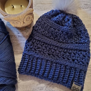 May include: A navy blue crocheted beanie with a faux fur pom-pom. The hat features a textured stitch pattern and a small metal tag. A skein of navy blue yarn and a candle in an owl-shaped holder are also visible.