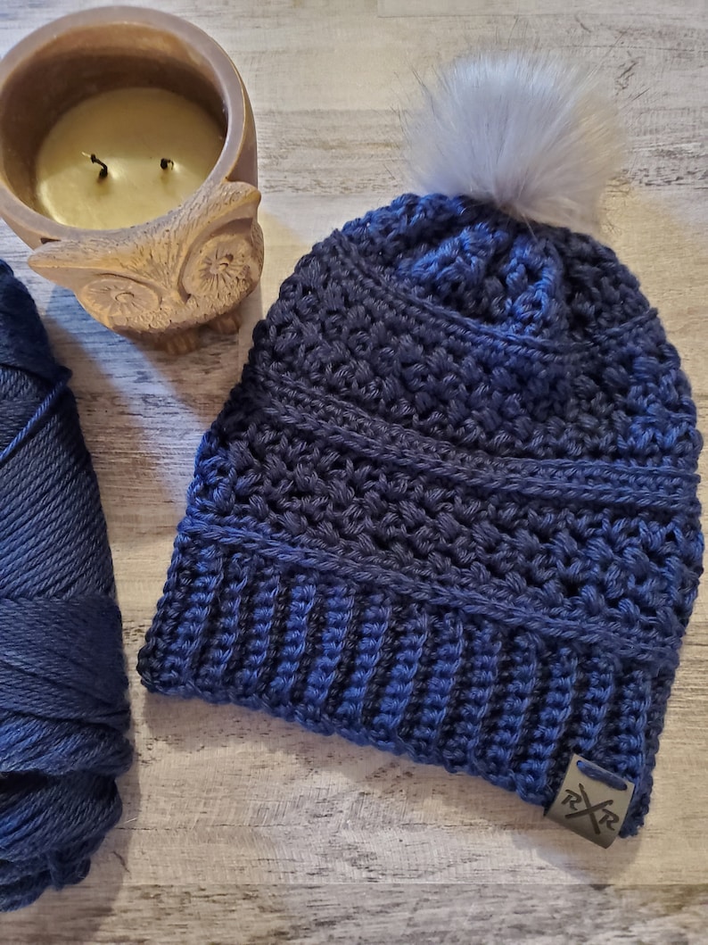 May include: A navy blue crocheted beanie with a faux fur pom-pom. The hat features a textured stitch pattern and a small metal tag. A skein of navy blue yarn and a candle in an owl-shaped holder are also visible.