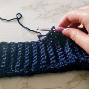 May include: A close-up of a navy blue crocheted hat in progress. The ribbed texture is visible, with a crochet hook inserted into the work. The yarn is a matching navy blue color, and the project is on a white and gray marble surface.