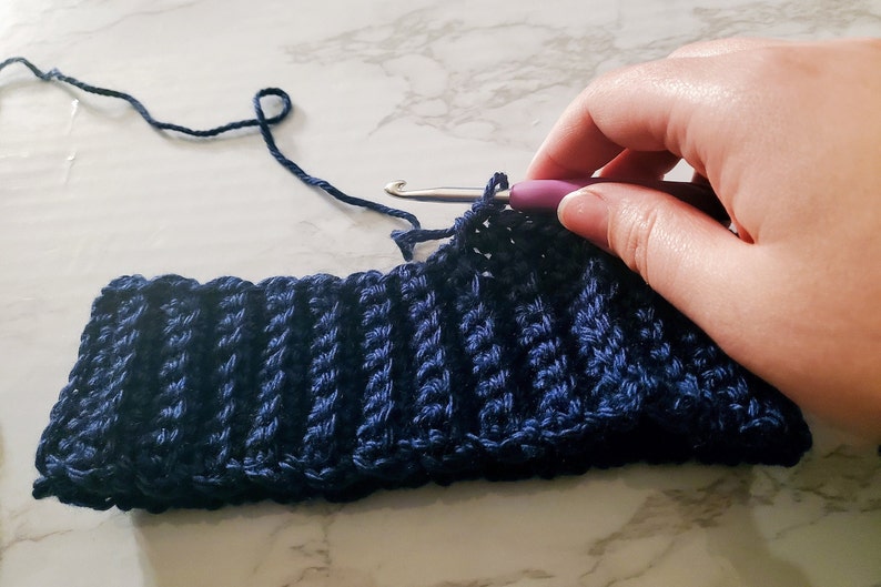 May include: A close-up of a navy blue crocheted hat in progress. The ribbed texture is visible, with a crochet hook inserted into the work. The yarn is a matching navy blue color, and the project is on a white and gray marble surface.