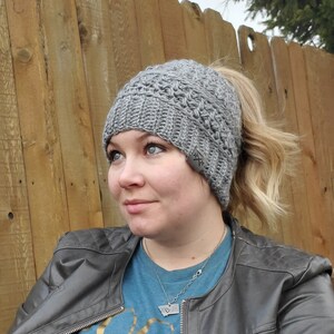 May include: A gray crocheted beanie with a ponytail opening. The hat has a textured pattern and a small tag on the side. The person is wearing a gray leather jacket and a blue shirt.